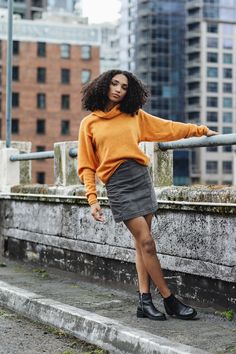 a woman in an orange sweater and grey skirt is leaning against a railing with her hand on the rail