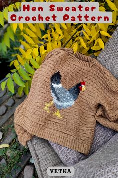 a sweater with a chicken on it sitting next to some yellow flowers and bushes in the background