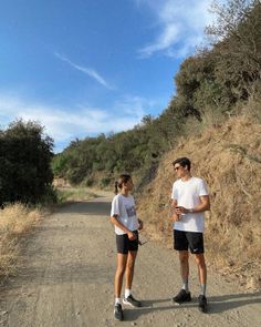 a man and woman standing on a dirt road next to a hill with trees in the background