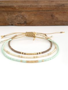 three different bracelets with gold beads and green cords on a white table next to a wooden box