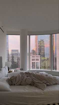 an unmade bed in front of two large windows with city skylines behind it