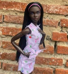 a doll with long black hair wearing a pink and white striped dress standing in front of a brick wall