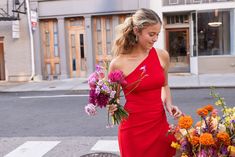 a woman in a red dress walking down the street with flowers on her hand and holding a bouquet