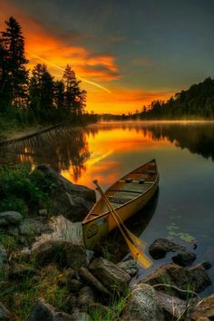 a canoe is sitting on the shore of a lake as the sun sets in the background