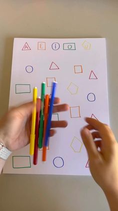 two hands holding colored pencils in front of a sheet of paper with geometric shapes on it