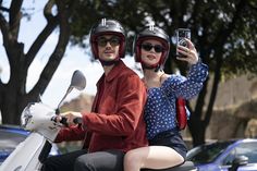 a man and woman riding on the back of a scooter while holding up their cell phones