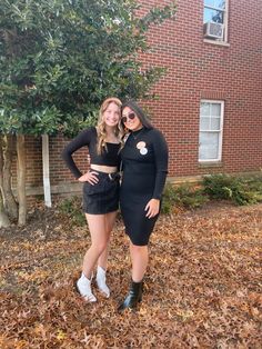 two women standing next to each other in front of a brick building with leaves on the ground