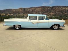 an old blue and white car parked in a lot with mountains in the back ground