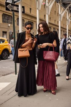 two women standing next to each other while looking at their cell phones on the street