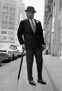 an old photo of a man in a suit and tie holding a cane on the street