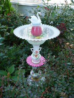 there is a bird bath in the middle of some bushes and plants with a cup on top