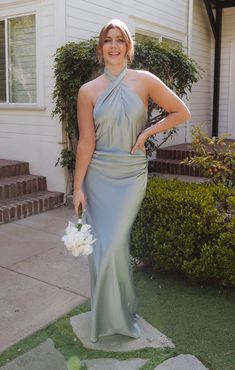 a woman standing in front of a house wearing a gray dress and holding a bouquet