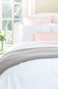a white bed with pink and grey pillows on it's headboard in front of a window
