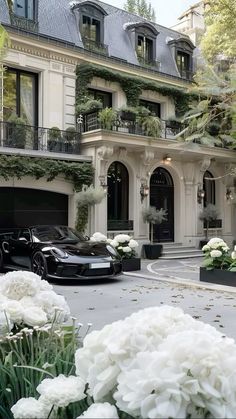 a black car parked in front of a large building with white flowers on the driveway