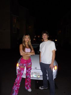 a man standing next to a woman in front of a car at night with her arms crossed
