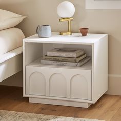 a nightstand with books and a lamp on top of it next to a white bed