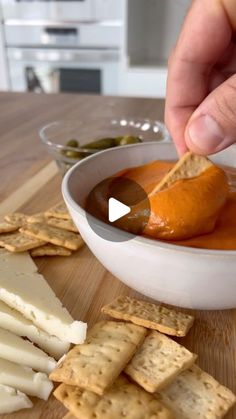 a person dipping sauce into crackers in a white bowl on a wooden cutting board