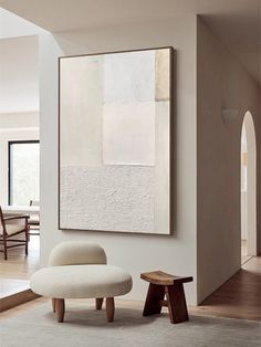 an abstract painting hangs on the wall next to a white chair and footstool