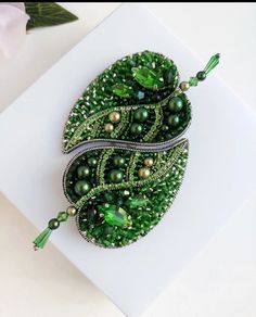 a green brooch with pearls and beads on it sitting on a white surface next to a pen