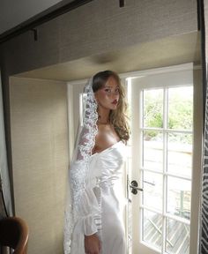 a woman in a white wedding dress standing by a door wearing a veil and looking at the camera