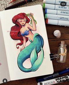 a drawing book with an image of a mermaid on it next to markers and pens