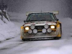 a rally car driving down a snowy road in the snow with its headlights turned on