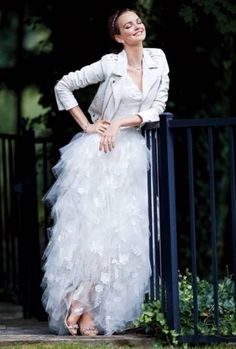 a woman in a white dress and jacket leaning on a fence with her hands on her hips