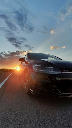 a black car driving down the road with the sun setting in the distance behind it