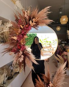 a woman standing in front of a mirror with dried plants on the wall behind her