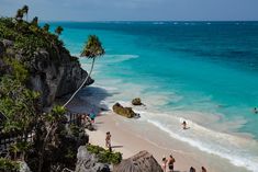 people are on the beach and in the water near some rocks, palm trees, and blue water