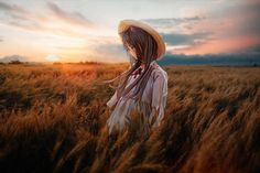 a woman with long hair standing in the middle of a wheat field at sunset wearing a straw hat