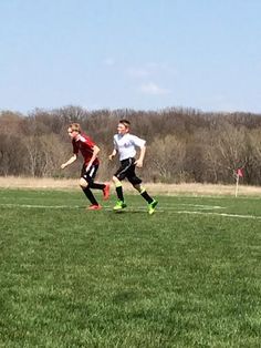 two young men playing soccer on a field with trees in the backgrouds