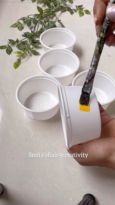 a person holding a paintbrush over four empty cups with plants in the background on a table