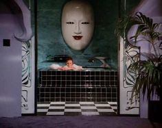 a woman in a bathtub with a mask on the wall above it and a potted plant next to it