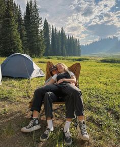 two people are sitting on a bench in the grass near tents and trees, while one person is sleeping