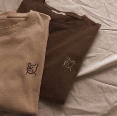 three t - shirts on a bed with white sheets and one has a teddy bear embroidered on it