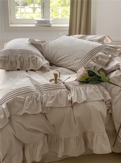 a bed with ruffled sheets and pillows in front of a window next to a flower