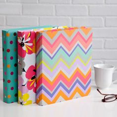 three colorful notebooks sitting next to each other on a white table with a cup