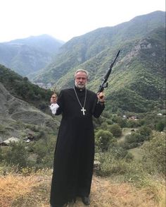 a priest holding two large metal objects in front of mountains and valleys with hills behind him