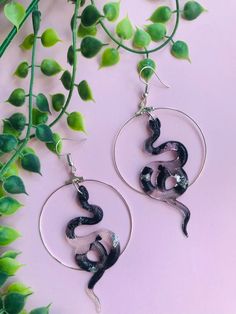 two black and white snakes are hanging from silver hoop earrings on a pink background with green leaves