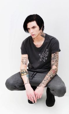 a man with tattoos sitting on the ground wearing grey pants and a black t - shirt