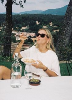 a woman sitting at an outdoor table eating food and drinking from a wine glass in front of her