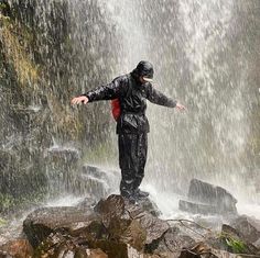 a man standing on top of a rock in front of a waterfall with his arms outstretched