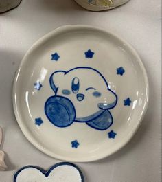 small to medium sized white porcelain plate , in the middle there is a handpainted Kirby. A character from nintendo, the kirby is encircled by 8 small stars Kirby Ceramic Mug, Kpop Pottery Ideas, Kuromi Pottery Painting, Cute Pottery Aesthetic, Porcelain Mug Painting, Porcelain Plate Design, Kirby Ashtray, Cute Ceramic Bowl Designs, Pottery Designs Plate