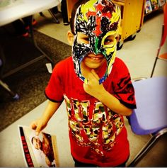 a young boy wearing a colorful mask and holding a book in front of his face