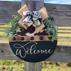 a welcome sign is hanging on a wooden fence with green plants and bow tie around it