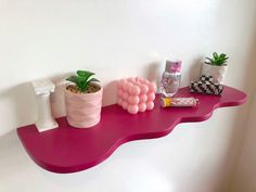 a pink shelf with some plants and other items on it next to a white wall