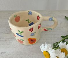 two daisies are next to a ceramic cup with fruit and berries on it sitting on a table