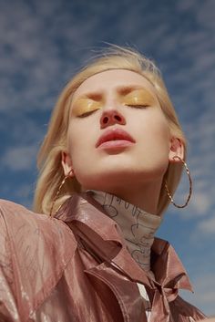 a woman with blonde hair and gold eyeshadow is looking up into the sky