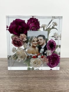 a glass frame with flowers and a couple in the center on a wooden table next to a white wall